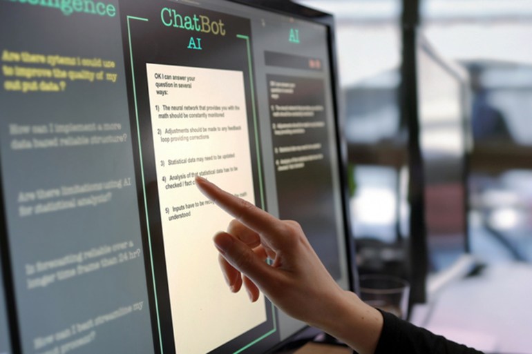 30-1694950065 Close-up stock photograph showing a touchscreen monitor being used in an open plan office. A woman’s hand is asking an AI chatbot pre-typed questions & the Artificial Intelligence website is answering.