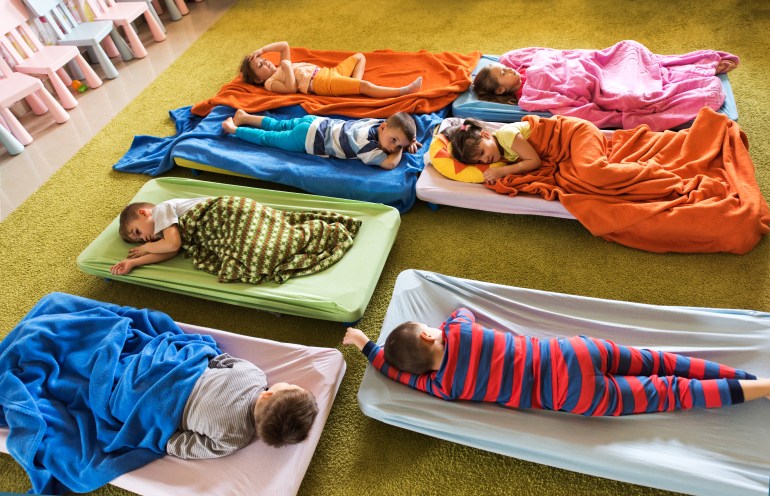 gettyimages-539673988-1706896618 High angle view of group of tired kids in pajamas resting on beds at kindergarten.