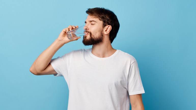 003-5-2 A man in a t-shirtشرب 8 أكواب من الماء يوميا.. حقيقة أم خرافة؟ and trousers on a blue background drinking water from a glass ; Shutterstock ID 1261170649; purchase_order: multimedia; job: ; client: ; other: