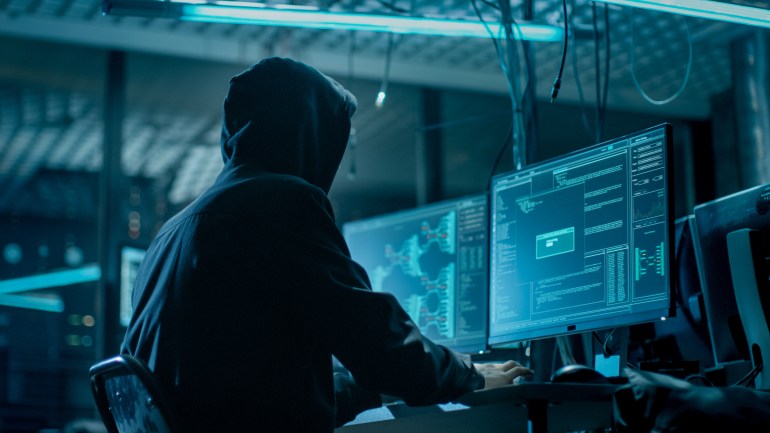 gettyimages-817486028-1695037118 Shot from the Back to Hooded Hacker Breaking into Corporate Data Servers from His Underground Hideout. Place Has Dark Atmosphere, Multiple Displays, Cables Everywhere.