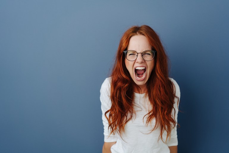 gettyimages-1251304747-1715173189 Angry young woman throwing a temper tantrum yelling at the camera with a furious expression over a blue studio background with copy space