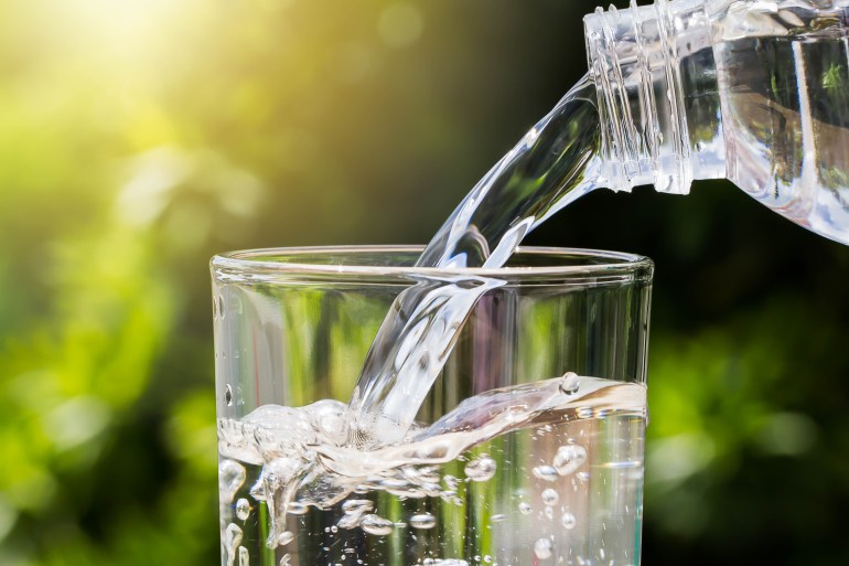 shutterstock_1036240708-1719680870 Drinking water pouring from bottle into glass on blurred green nature background