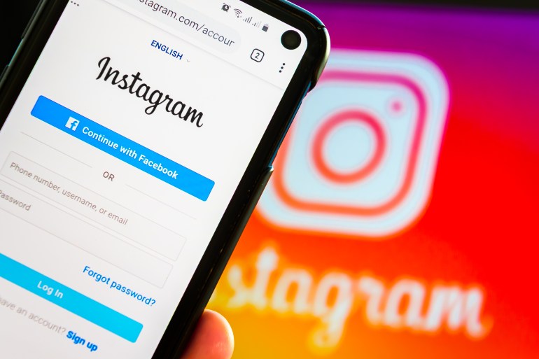 shutterstock_1714022380-1697355544 Closeup Of A Hand Holding Mobile Smart Cell Phone Device With Instagram Social Network Log In Screen Over a Notebook With Instagram Web Image On Display
