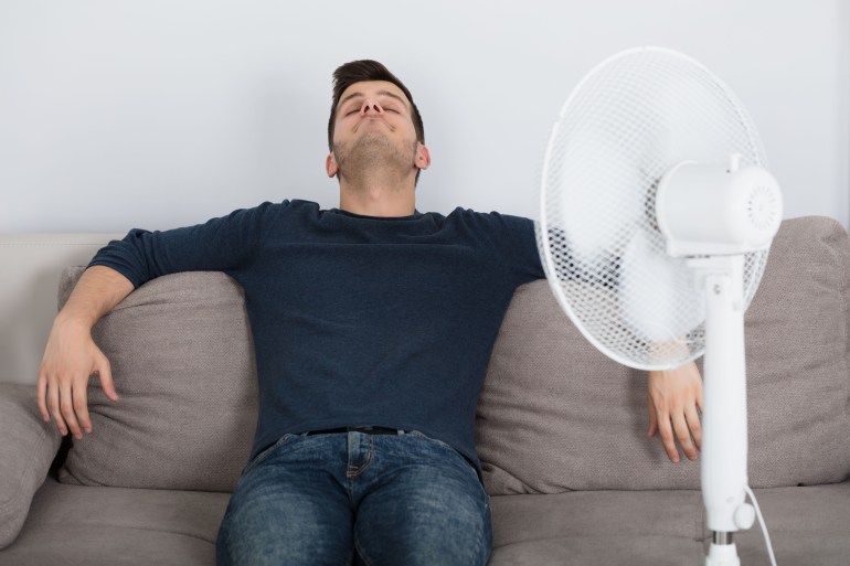 shutterstock_734181577-1722091935 Young Man Sitting On Couch Cooling Off With Fan During Hot Weather At Home; Shutterstock ID 734181577; purchase_order: ajanet; job: ; client: ; other: