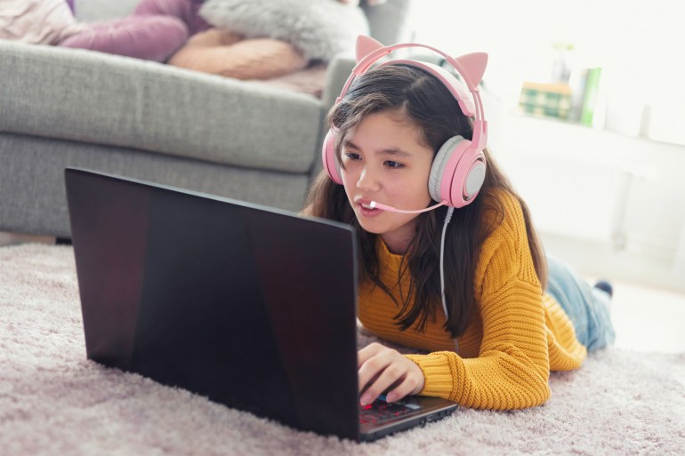 2323232 A girl laying down on a living room carpet and playing an online video game on a laptop computer while communicating using a headset with microphone.