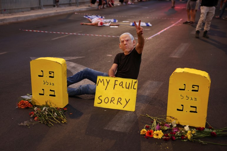 A person wearing a mask depicting Israeli Prime Minister Benjamin Netanyahu sits with a placard, as people demonstrate against Netanyahu's government and call for the release of hostages in Gaza, amid the Israel-Hamas conflict, in Tel Aviv, Israel, August 17, 2024. REUTERS/Florion Goga