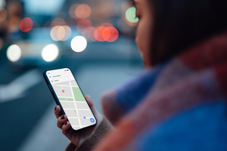 gettyimages-1306359673-1723720481 Over the shoulder view of woman using GPS navigation app on smartphone to navigate and search for location in city. Travel and navigation concept. Hand holding mobile phone mockup. غيتي