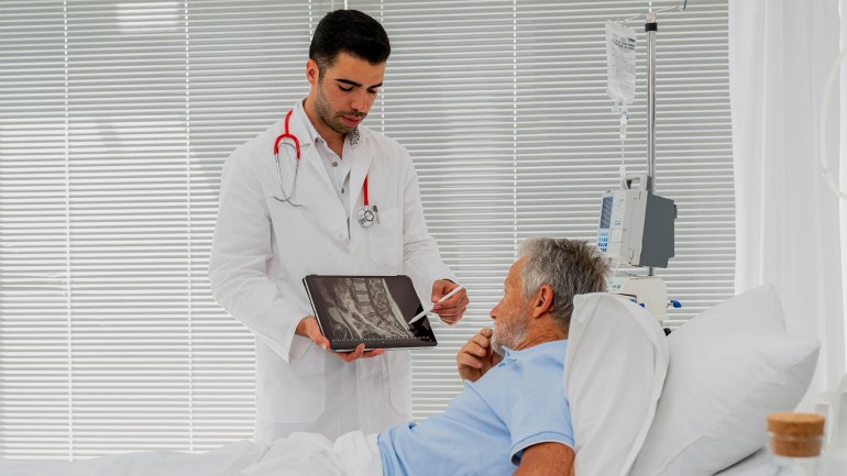 gettyimages-1400863470 غلاف فيديو بعنوان: هل هشاشة العظام حكر على النساء؟ This is why you're feeling pain - stock photo Hospital Ward: Friendly Doctor Talks to Senior male Patient Resting in Bed, Young Doctor using a Digital Tablet during a Consultation with a Senior Man GettyImages-1400863470