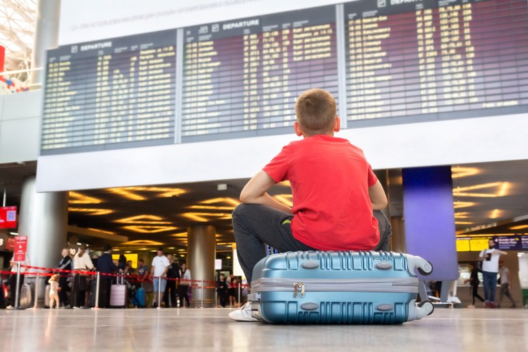 gettyimages-1662197206-1723451292 Lonely child sitting on a suitcase and waiting for departure at the airport looking at the arrivals board.