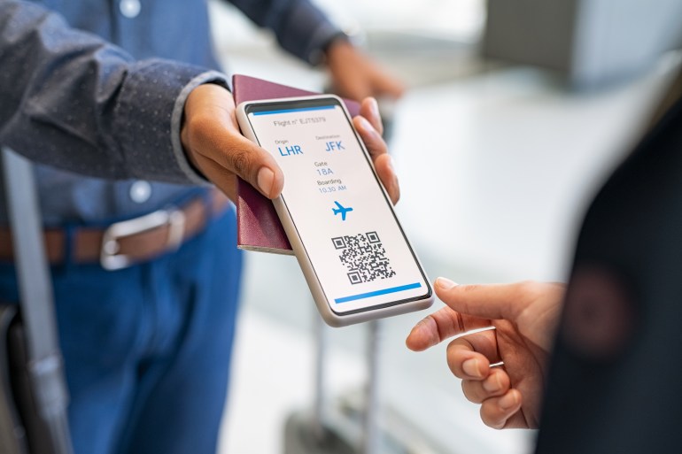 shutterstock_2099982607-1724784911 Close up hand of man showing electronic boarding pass to flight attendant on phone. Hostess checking electronic flight ticket at boarding gate. Check in counter and online air ticket at airport. شترستوك