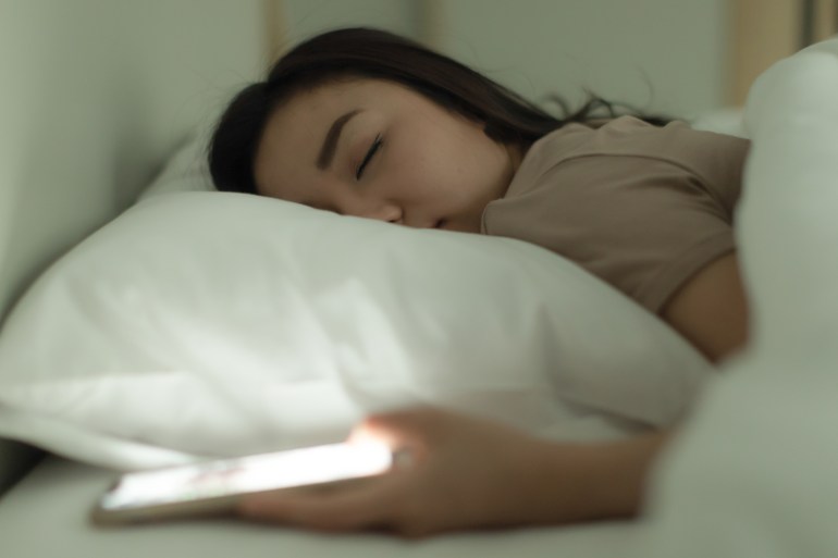 sxsxs2-1723125288 Cute young Asian woman laying on the bed while holding the switch on an open light smartphone in hand and asleep, nap with tired and deep sleep. Social media addict concept; Shutterstock ID 2176356905; purchase_order: aljazeera ; job: ; client: ; other: