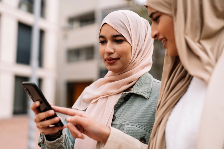 gettyimages-1308966202-1724597058 Portrait of two Arab female friends using a smartphone together while standing outdoors on the street.