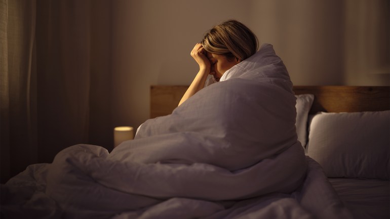 لماذا نتحدث مع أنفسنا؟ وما هو الاكتئاب المبتسم؟ Young woman holding her head in pain while being wrapped in a duvet on a bed in bedroom. Photographed in medium format.