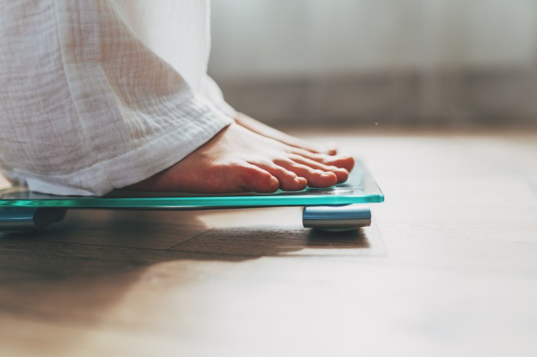 gettyimages-1500448393-1725200861 Female feet standing on electronic scales for weight control on wooden background. The concept of slimming and weight loss