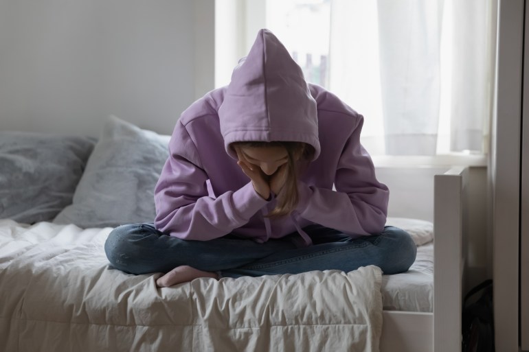 dep3-1719739793 Unhappy depressed teenage girl wearing sweatshirt sitting on bed alone, feeling lonely and misunderstood, stressed sad teenager thinking about troubles, hiding face, child and psychological problem; Shutterstock ID 2170756605; purchase_order: aljazeera ; job: ; client: ; other: