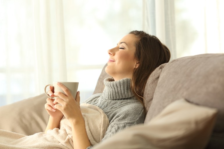 gettyimages-877068336-1731435912 Woman relaxing holding a coffee mug sitting on a sofa in the living room in a house interior