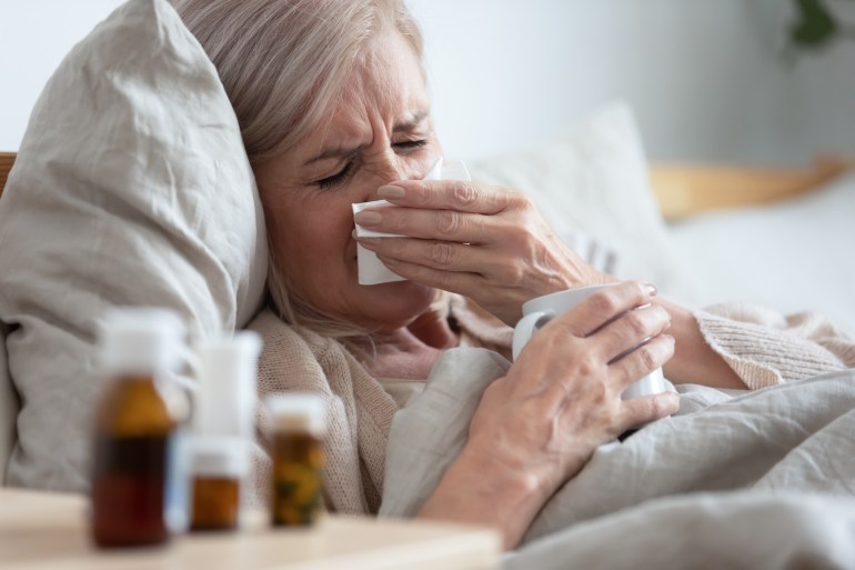 shutterstock_1536338474 Ill sick middle aged woman sneezing blowing running nose holding tissue sit on bed, upset old mature lady caught cold got flu influenza grippe symptoms drink hot tea taking medications at home alone