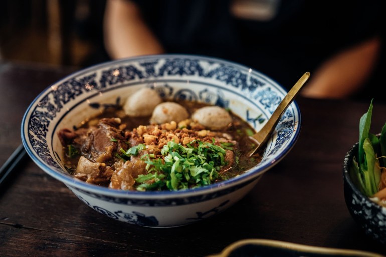 23123165444-1734888427 Close-up shot of Chinese beef noodle soup in decorative ceramic bowl, freshly served on a wooden table. Asian food cultures.