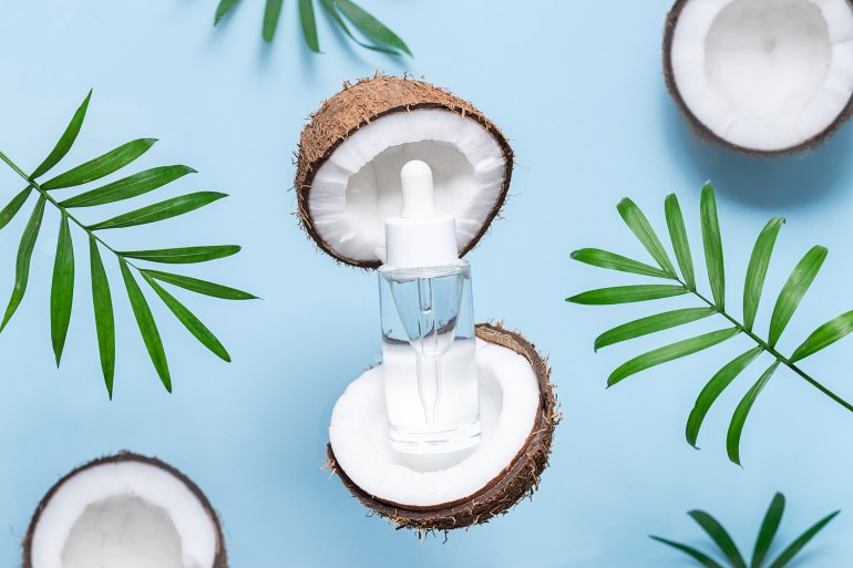 shutterstock_2388842701-1734021231 Skin care serum in glass bottle with dropper on light blue background with palm leaves and fresh aromatic coconuts. Natural cosmetic with coconut oil. Organic skin care products.