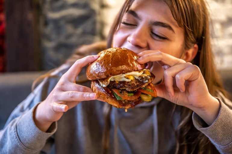 gettyimages-1318483615-1718040820 Close-up of a girl eating a big burger with meat and vegetables on a blurred background.