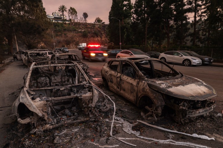 rr-3-1736574147 Burned-out cars by the Palisades Fire are seen pushed out of the way by firefighters in the Pacific Palisades neighborhood of Los Angeles, California on January 10, 2025. Massive wildfires that engulfed whole neighborhoods and displaced thousands in Los Angeles have killed at least 10 people, authorities said, as California's National Guard soldiers readied to hit the streets to help quell disorder.