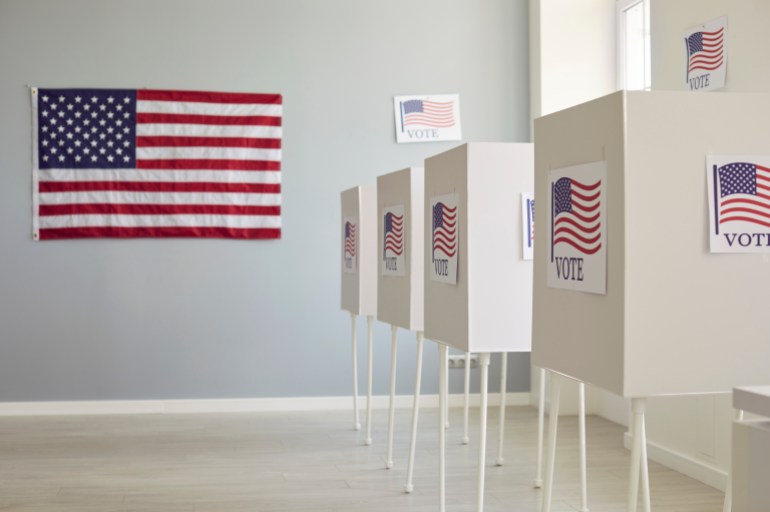 shutterstock_2475726979-1728484169 White empty voting booths at polling station with American flag. Interior of vote center ready for presidential elections in the United States. Democracy and election day concept.