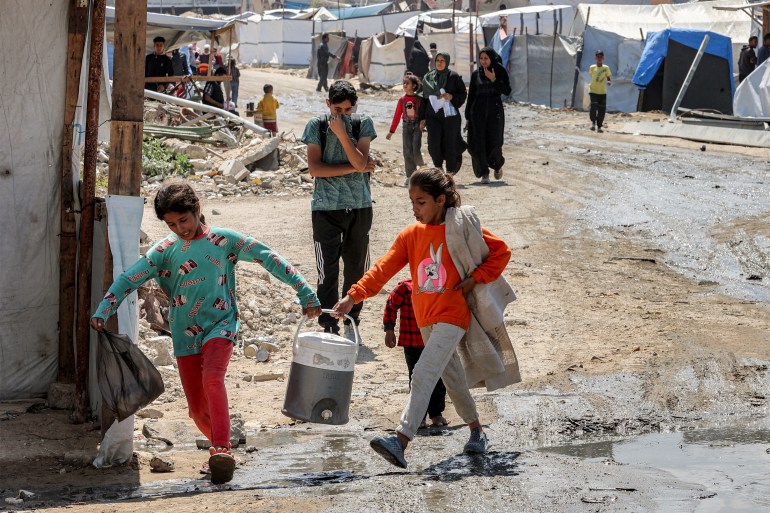 afp_68077e27a61e-1745321511 Two girls carry together a water cooler as they walk past tent shelters in the Nasr neighbourhood in western Gaza City on April 15, 2025.