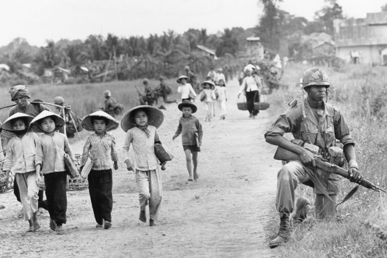 ap955927196003-1745930753 Vietnam War U.S. Vietnamese Civilians In an area heavily infiltrated by Viet Cong, a U.S. 1st Division soldier guards Route 7 as Vietnamese market women and schoolchildren return home to the village of Xuan Dien from Ben Cat, December 1965. (AP Photo/Horst Faas)
