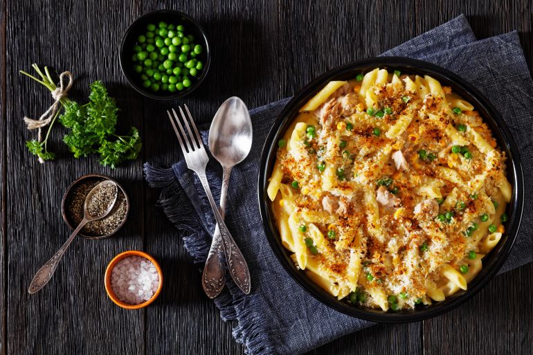 shutterstock_2230398657-1745602135 تونة Tuna Mornay, Creamy Tuna Casserole Penne Pasta Bake in baking dish on dark wooden table with fork and spoon, horizontal view from above, flat lay