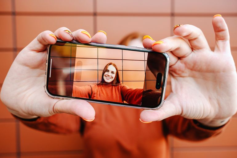 Happy woman taking selfie through smart phone in front of brown wall