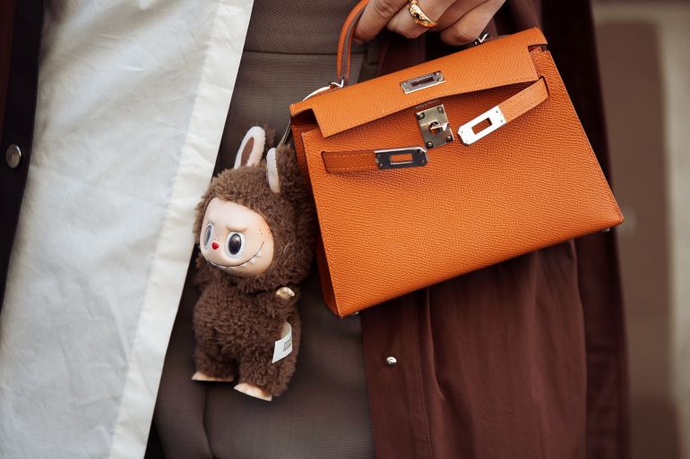 56tyer-1747322152 PARIS, FRANCE - MARCH 08: A guest wears brown coat, white blouse, orange Hermes bag with a bag charm brown Labubu monster, grey pants outside the Hermes fashion show during the Womenswear Fall/Winter 2025/2026 as part of Paris Fashion Week on March 08, 2025 in Paris, France. (Photo by Raimonda Kulikauskiene/Getty Images)