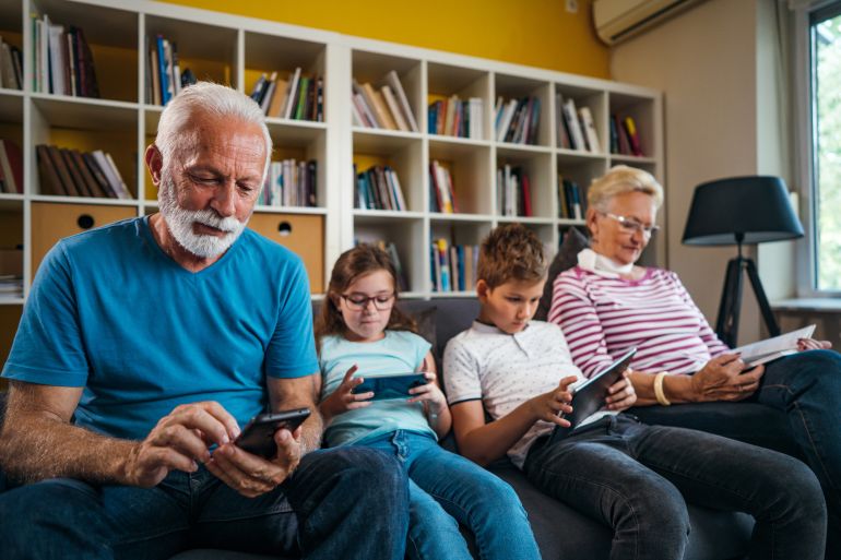 Senior man and kids are using wireless technology while relaxing in the living room. Senior woman is reading a book.