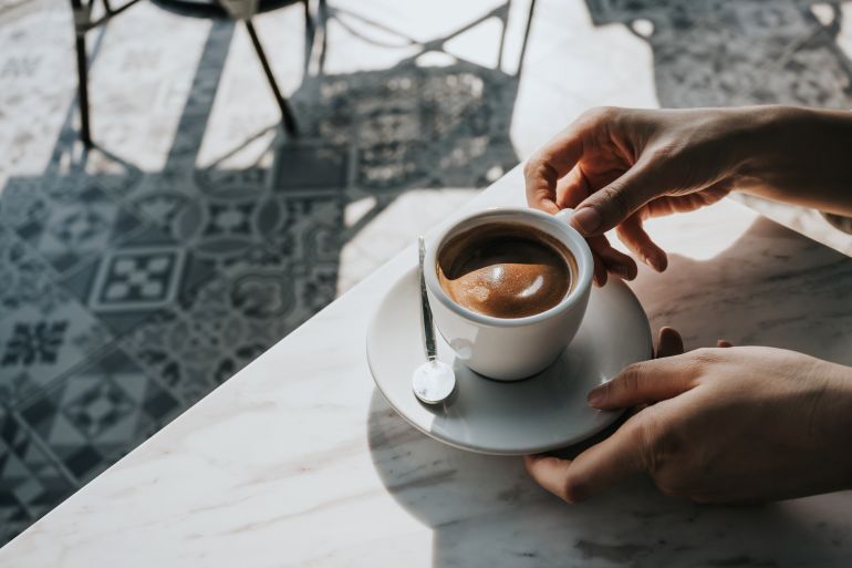 gettyimages-1357844240-1746200898 Close up of woman's hand holding a cup of coffee, drinking coffee in outdoor cafe against beautiful sunlight, having a relaxing moment. Enjoying life's simple pleasures