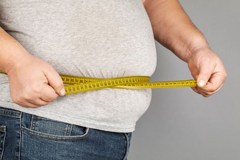 shutterstock_1372581767-1747928489 A man measures his fat belly with a measuring tape. on a gray background