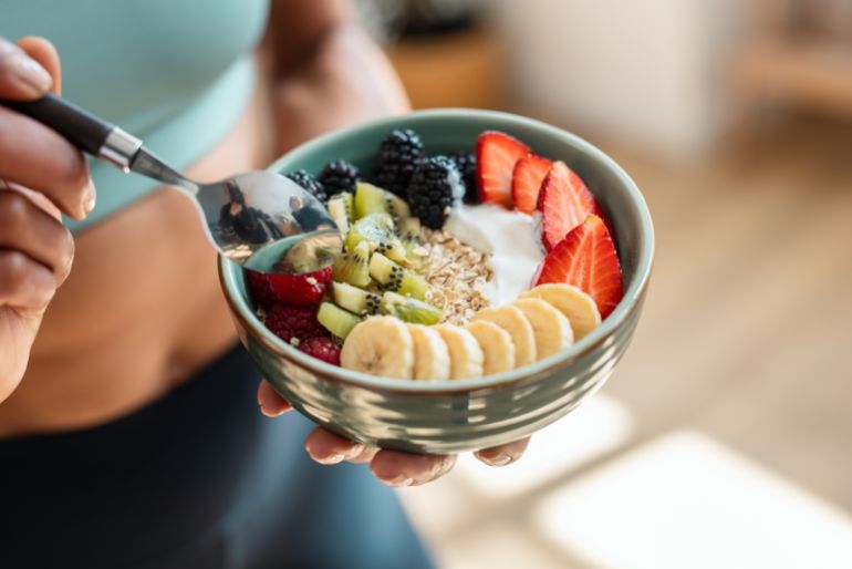 shutterstock_2470596591-1761746237 Close up of athletic woman eating a healthy fruit bowl in the kitchen at home
