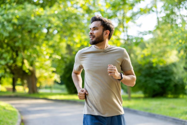 shutterstock_2519195487-1761746304 Man running in park surrounded by lush greenery showing joy and determination during outdoor workout wearing fitness tracker promoting health and wellness