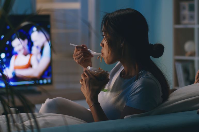 65uyh-1762186749 Asian woman eating takeaway noodles and watching TV while sitting on couch and having dinner in evening at home