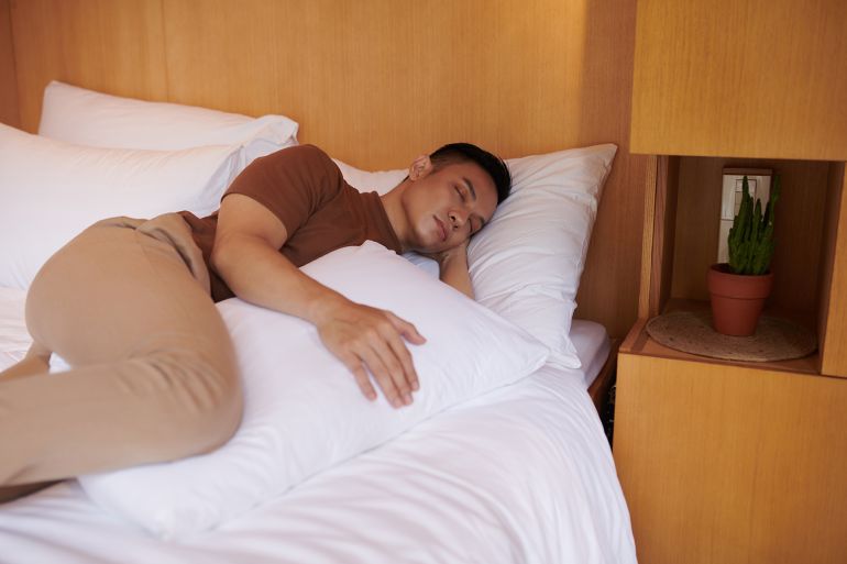 Young man sleeping with pillow between legs, which keeps his pelvis neutral and prevents spine from rotating during night by DragonImages ادوبي ستوك