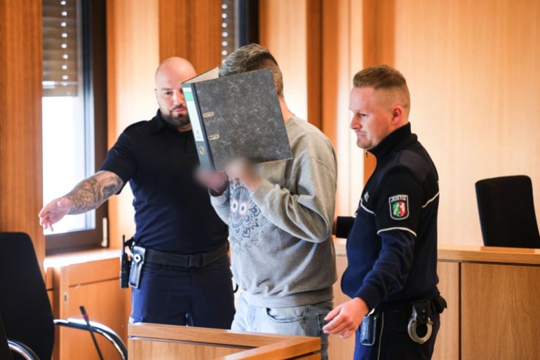 afp_690b894232a8-1762363714 Picture taken on March 24, 2025 shows the defendant (C), a former nurse in a palliative care unit, hiding his face behind a folder as he is brought to court for the start of his trial in Aachen, western Germany.