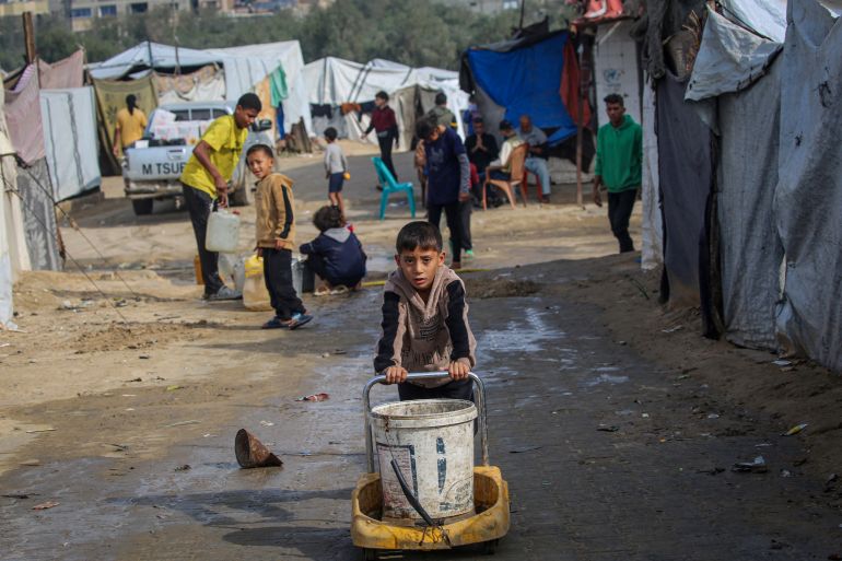 afp_692c031f63b6-1764492063 A boy pushes a bucket as children fill their containers with water at the Nuseirat camp for displaced Palestinians in the central Gaza Strip on November 29, 2025.