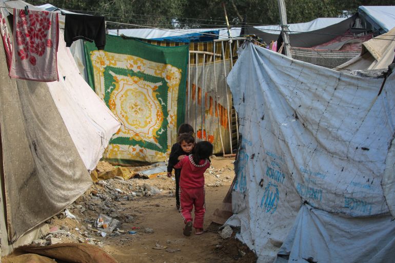 afp_692c032760e6-1764492071 A girl carries a child as they walk between makeshift shelters at the Nuseirat camp for displaced Palestinians in the central Gaza Strip on November 29, 2025.