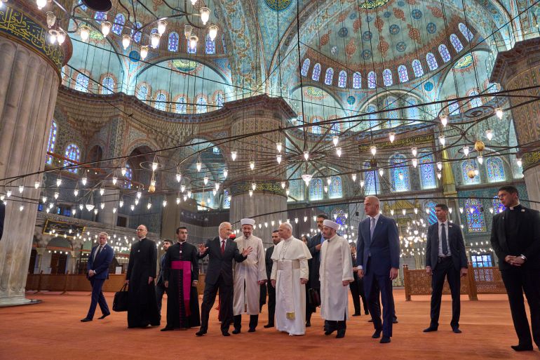 ap_692b0282c385e-1764426370 Pope Leo XIV, center, walks with Muezzin Musa Asgın Tunca, fourth from left, Istanbul's Mufti Emrullah Tuncel, fifth from left, the Imam of Mosque Sultanahmet Fatih Kaya, seventh from left, and Turkish Minister for Culture and Tourism Mehmet Nuri Ersoy, eighth from left, during a visit of the Sultan Ahmed Mosque in Istanbul, Saturday, Nov. 29, 2025. (AP Photo/Domenico Stinellis)