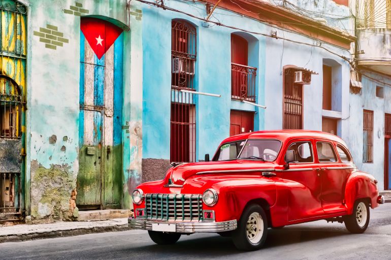 shutterstock_1176936922-1762085071 HAVANA,CUBA - SEPTEMBER 5,2018 : Classic car and and old building with the cuban flag in Old Havana