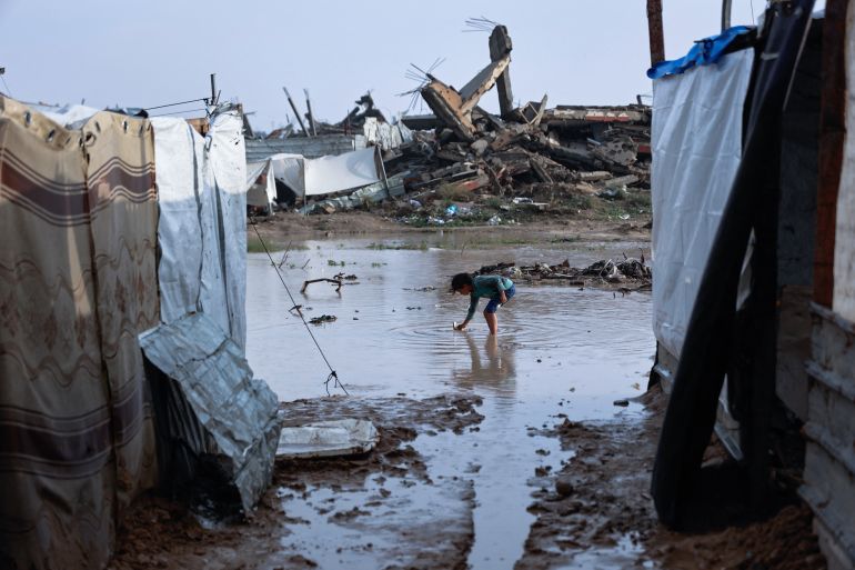 afp_693d06aed93c-1765607086 A girl stands in a pool of water at a makeshift camp sheltering displaced Palestinians after heavy rains in the Zeitoun neighbourhood of Gaza City on December 11, 2025.