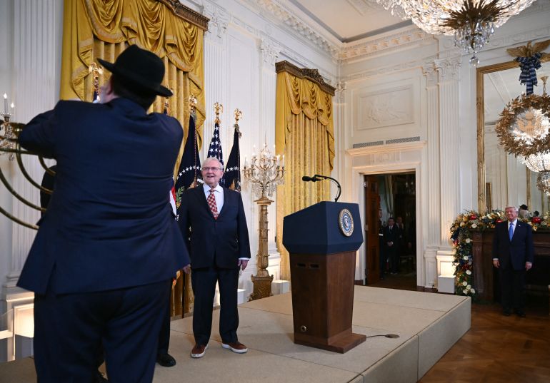 afp_69478669eeec-1766295145 (L-R) Rabbi Levi Shemtov lights the menorah as Holocaust survivor Michael Bornstein (C) and US President Donald Trump (R) look on during a Hanukkah reception in the East Room of the White House in Washington, DC, on December 16, 2025.