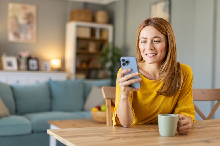 A joyful woman smiles while using her smartphone at a desk, capturing the essence of happiness, technology, and the comforts of home.