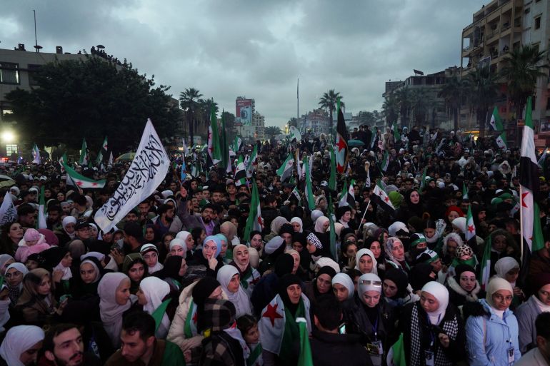 reuters_6937e1b9-1765269945 Syrians holds flags as they gather to mark the first anniversary of Bashar al-Assad's fall, in Latakia, Syria December 8, 2025. REUTERS/Karam al-Masri