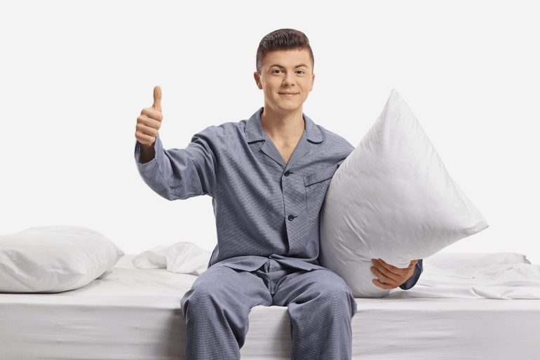 Teenager in pajamas seated on a bed holding a pillow and making a thumb up gesture isolated on white background