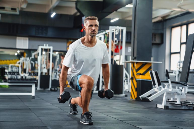 Young attractive sportsman doing split squats with weights in gym