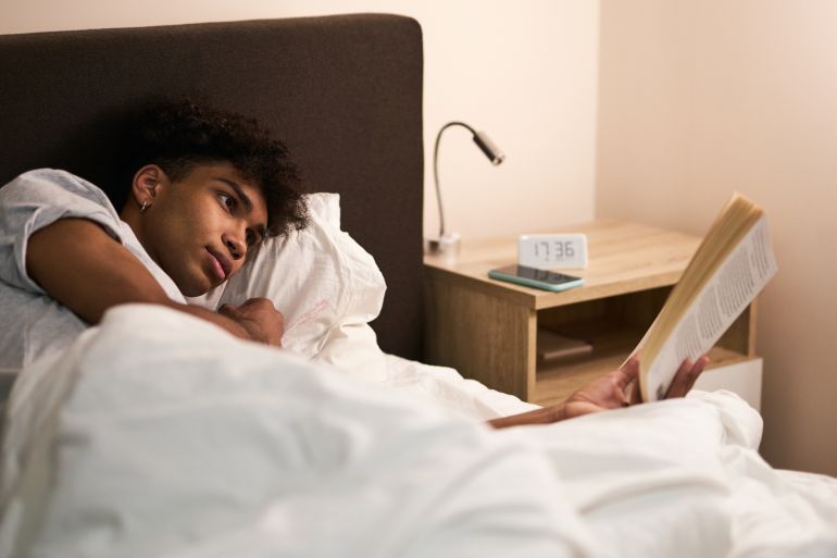 Young guy lying in the bed at home, resting and reading an interesting book before going to sleep